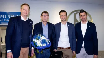 Vier Personen in Businesskleidung posieren vor einem S04‑Logo im Innenbereich; eine Person hält einen blau‑weißen Blumenstrauß, eine weitere eine Geschenkbox, rechts ein Schild mit „Museum“.