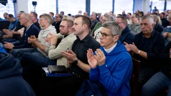 Reihen sitzender Personen bei einer Veranstaltung, alle klatschen; im Vordergrund eine Person im blauen Hoodie mit Notizbuch auf dem Schoß, unscharfer Hintergrund mit Publikum.