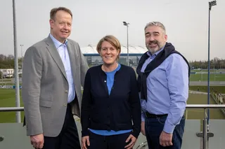 Michael Kalthoff, Christina Rühl-Hamers und Hürgen Gerdes stehen auf einem Balkon. Im Hintergrund das Trainingsgelände des FC Schalke 04 und die VELTINS-Arena.