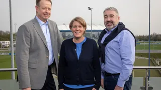 Michael Kalthoff, Christina Rühl-Hamers und Hürgen Gerdes stehen auf einem Balkon. Im Hintergrund das Trainingsgelände des FC Schalke 04 und die VELTINS-Arena.