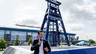 Matthias Tillmann mit Mikrofon vor dem blauen Förderturm‑Denkmal der Auf Schalke eG; im Hintergrund die VELTINS‑Arena und Besucher auf dem Vorplatz.